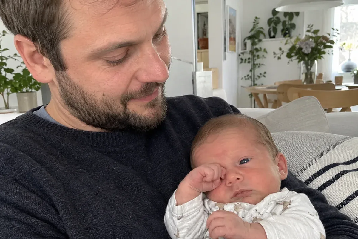 A 3 week old in a parent's arms rubbing one eye and staring past the camera