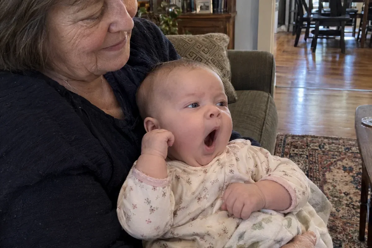 A 6 month old held in a parent's arms, rubbing one eye and staring off to the side