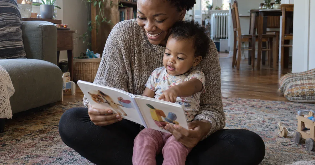 A Black mother with short natural hair sits on a patterned rug reading a board book with her 1 year old in her lap pointing at the pictures