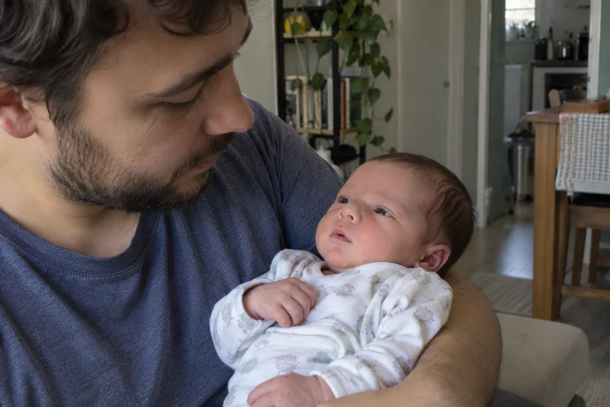A newborn lying awake in a parent's arms, staring off past the parent's face with a still, quiet look