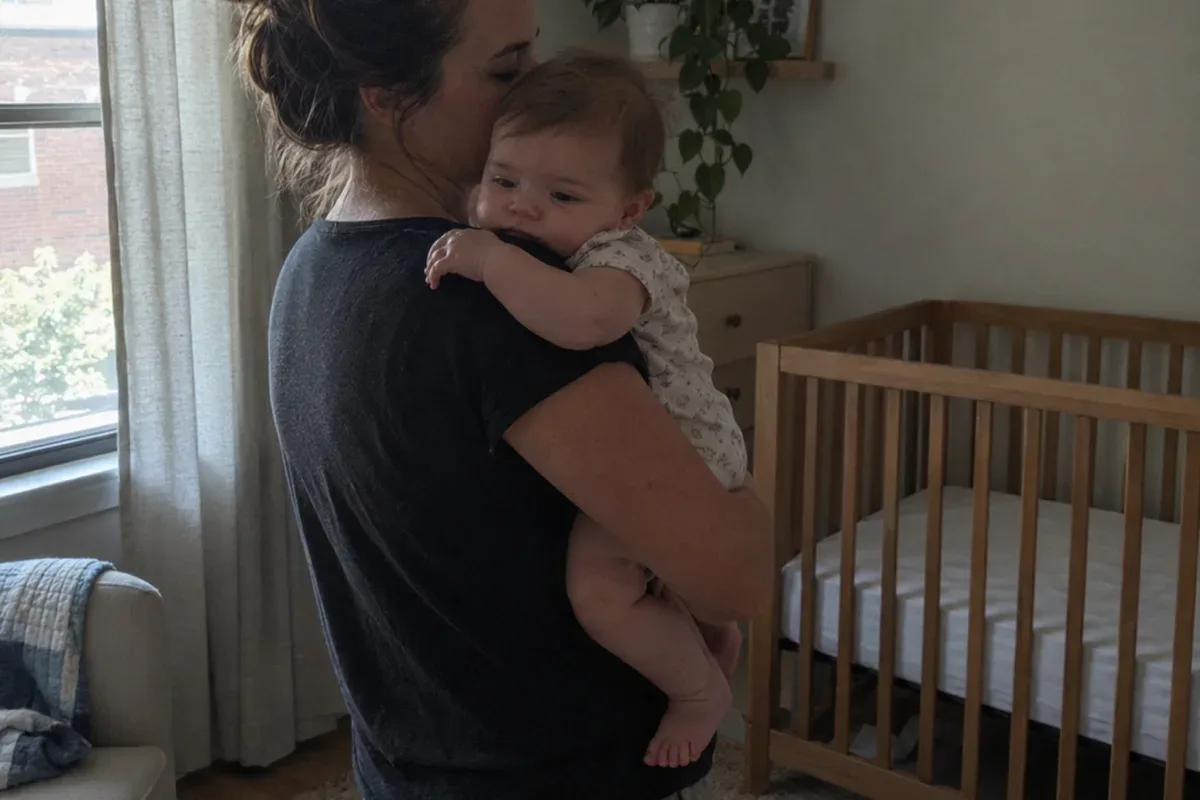 A parent sways in a dim nursery holding a drowsy 10 month old against their shoulder, an empty crib behind