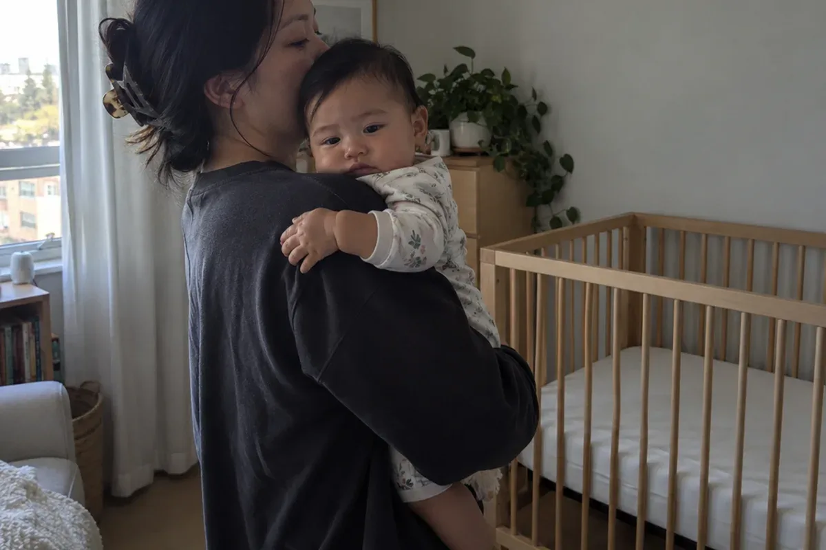 A parent sways in a dim nursery with a drowsy, still-awake 9 month old on their shoulder, empty crib in the background