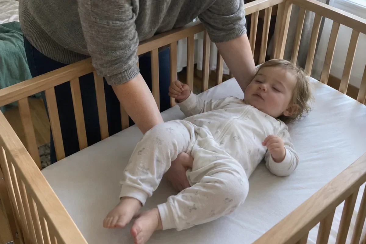 A parent's arms lower a drowsy 1 year old onto their back in an empty crib with only a fitted sheet