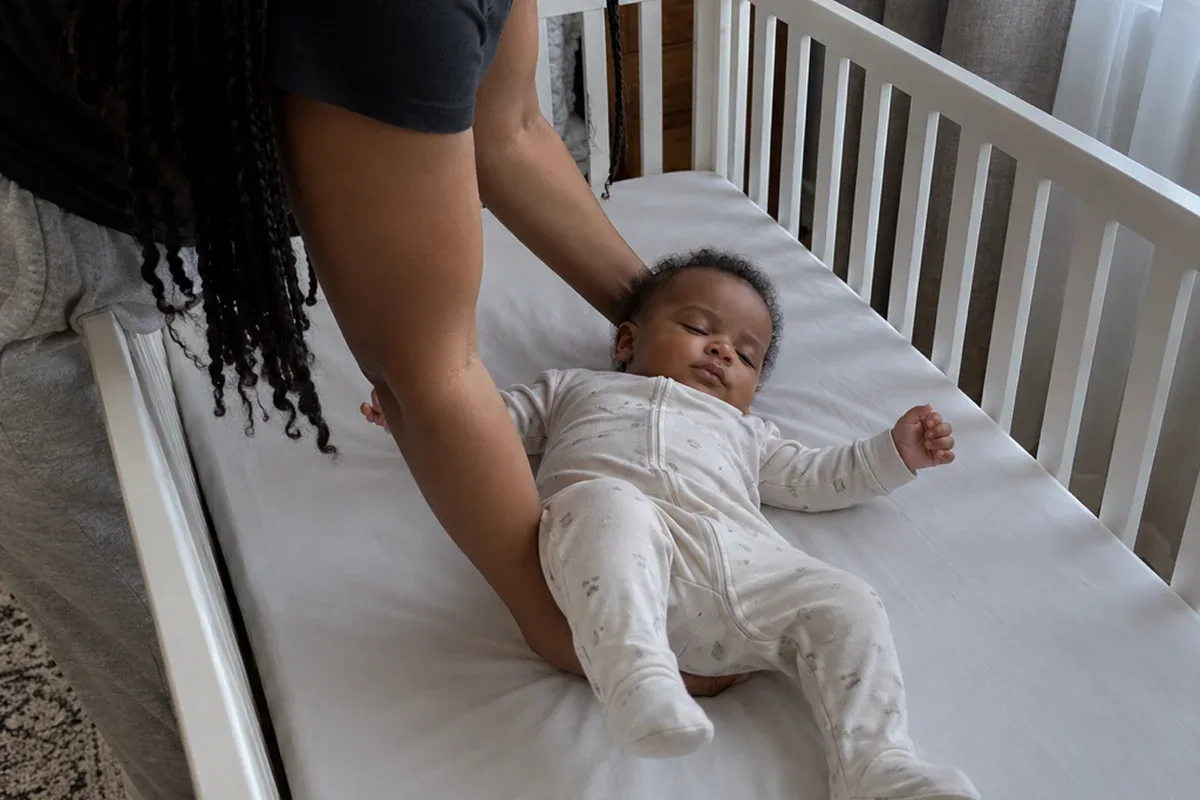 A parent's arms lower a drowsy 8 month old onto their back in an empty crib with a fitted sheet only