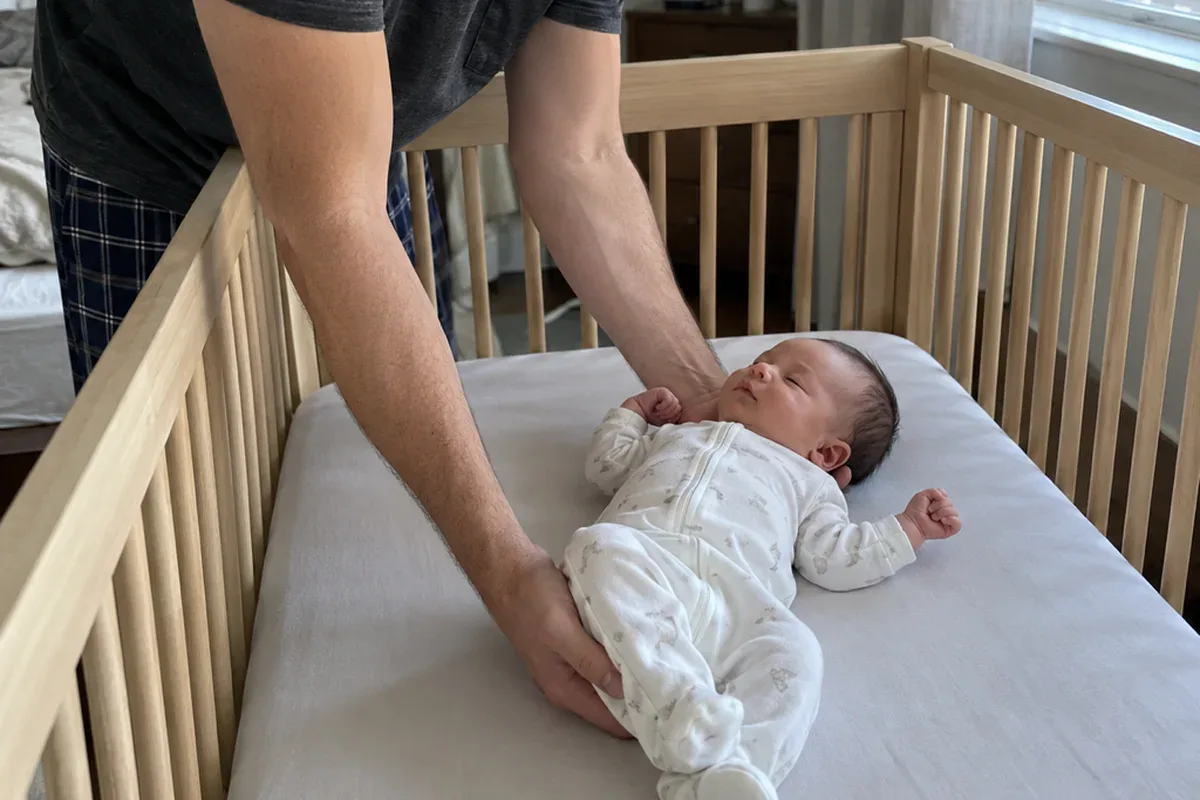 A parent's arms lowering a drowsy 1 month old onto their back in an empty crib with only a fitted sheet
