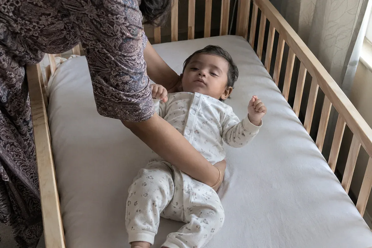 A parent's arms lowering a drowsy 10 month old onto their back in an empty crib with a plain fitted sheet