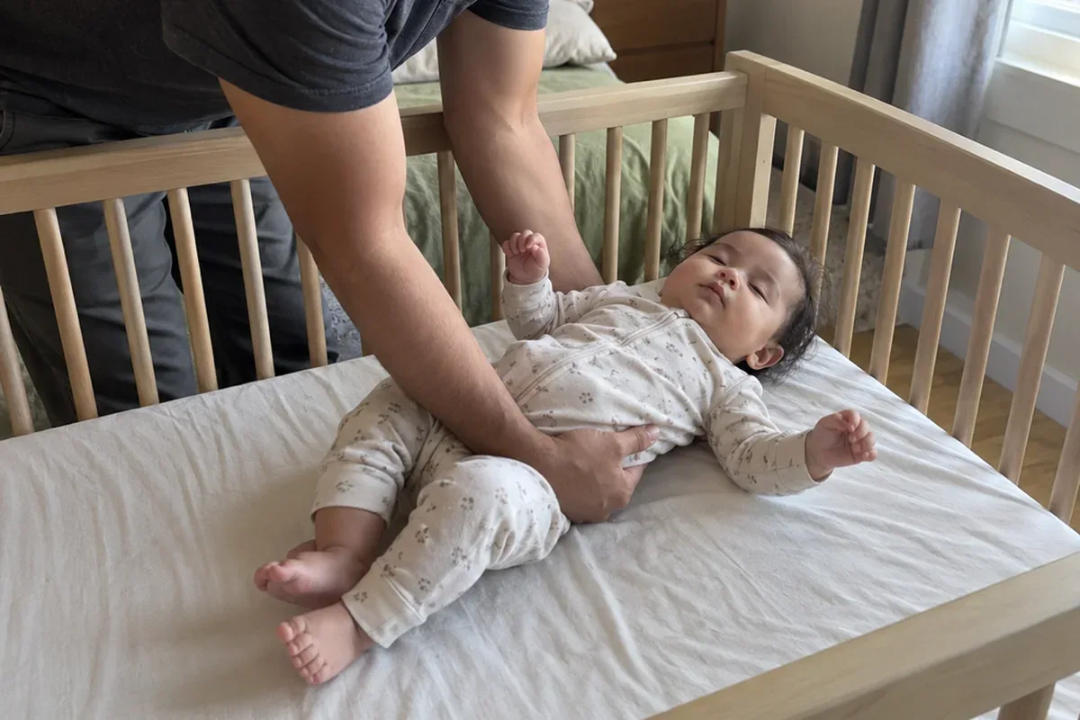 A parent's arms lowering a drowsy 11 month old onto their back in a bare crib with a fitted sheet