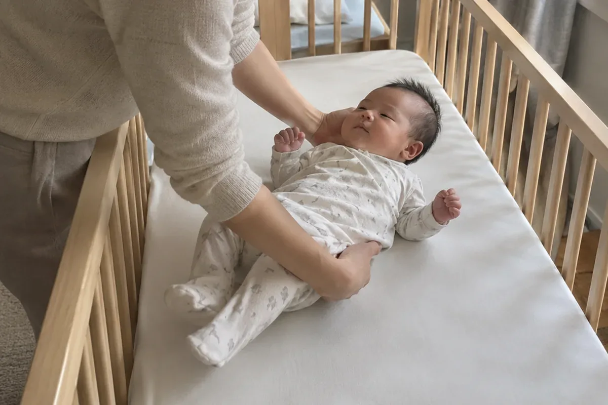 A parent's arms lowering a drowsy 2 month old onto their back in an empty crib with only a fitted sheet
