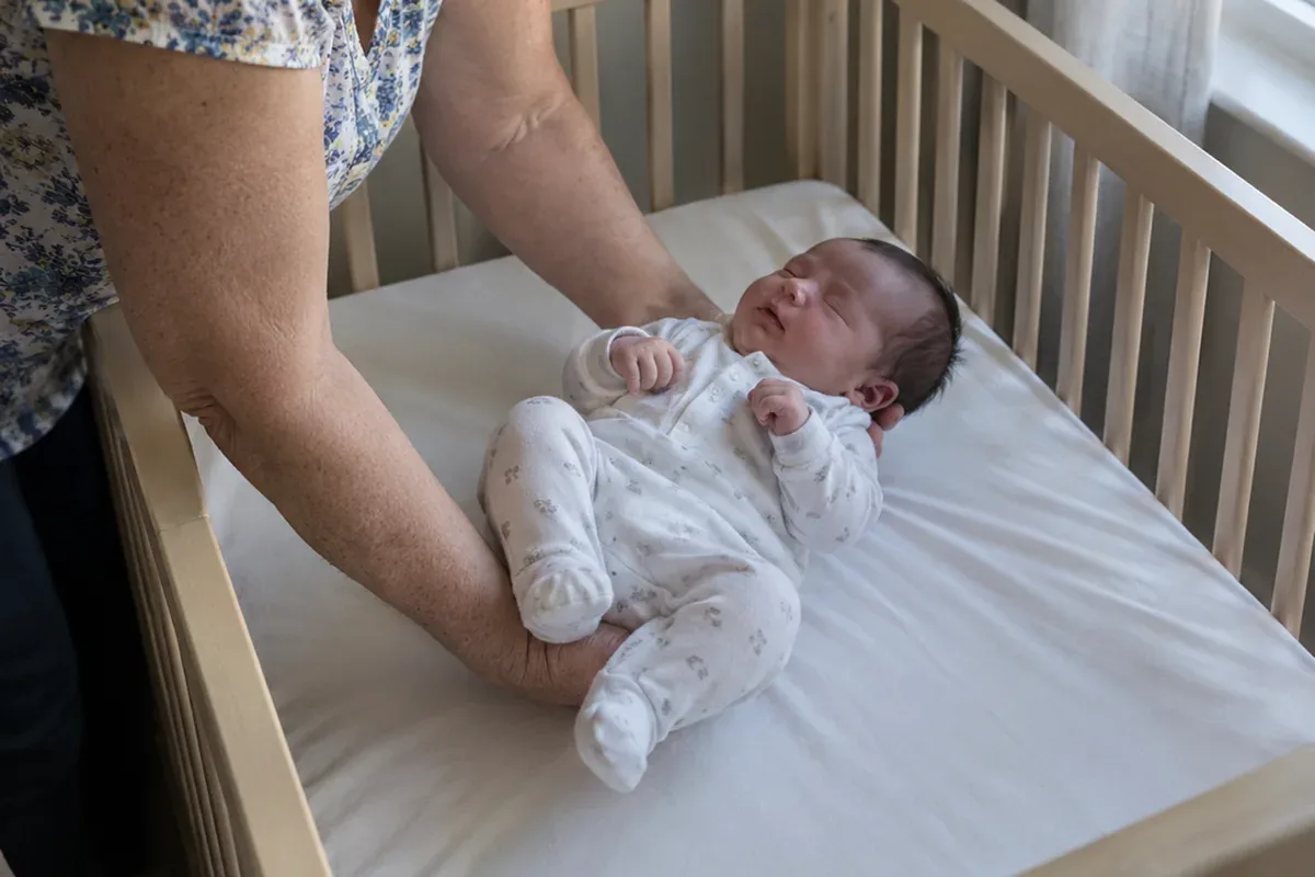 A parent's arms lowering a drowsy 3 week old onto their back in an empty crib with a fitted sheet only