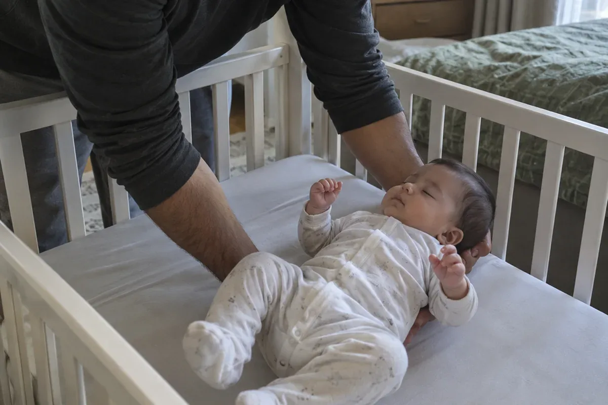 A parent's arms lowering a drowsy 4 month old onto their back in a bare crib with a fitted sheet only
