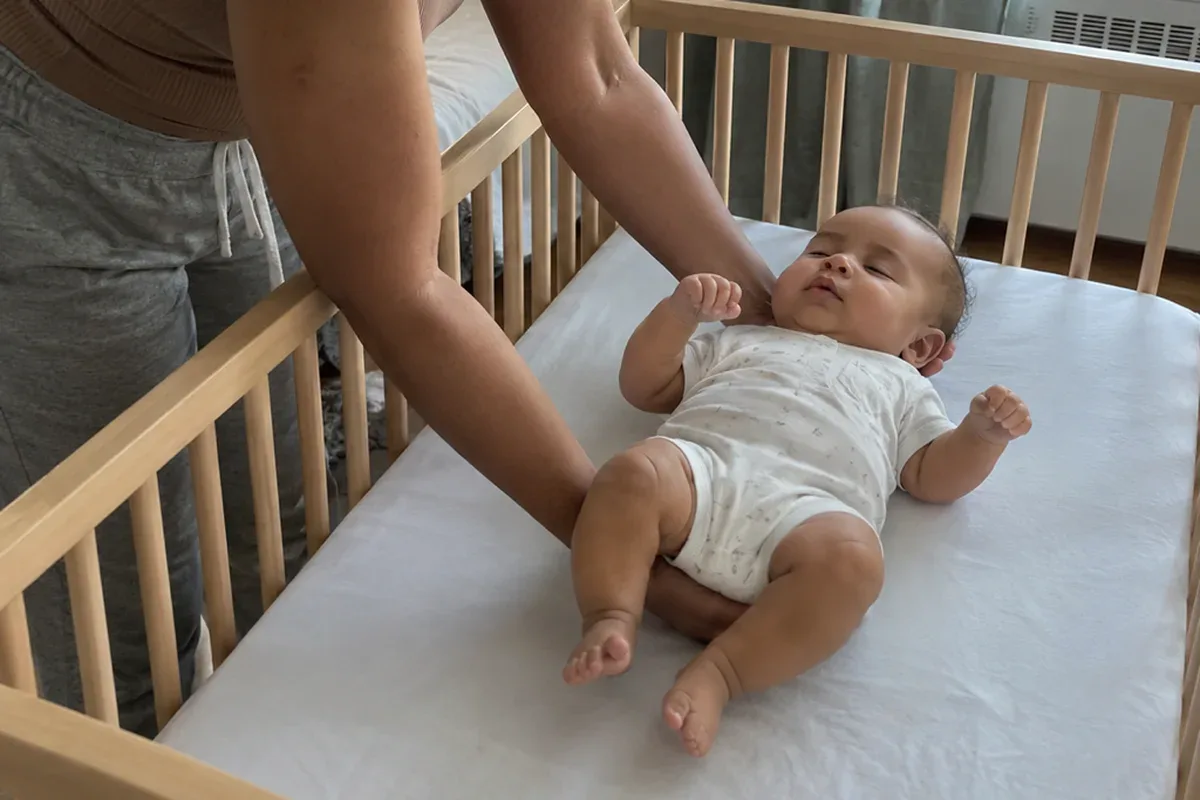 A parent's arms lowering a drowsy 5 month old onto their back in an empty crib with a fitted sheet