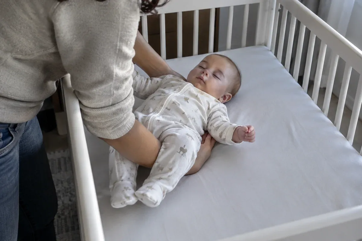 A parent's arms lowering a drowsy 6 month old onto their back in an empty crib that has only a fitted sheet