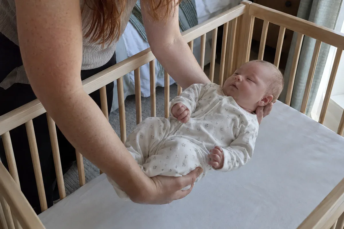 A parent's arms lowering a drowsy 6 week old onto their back in an empty crib with only a fitted sheet