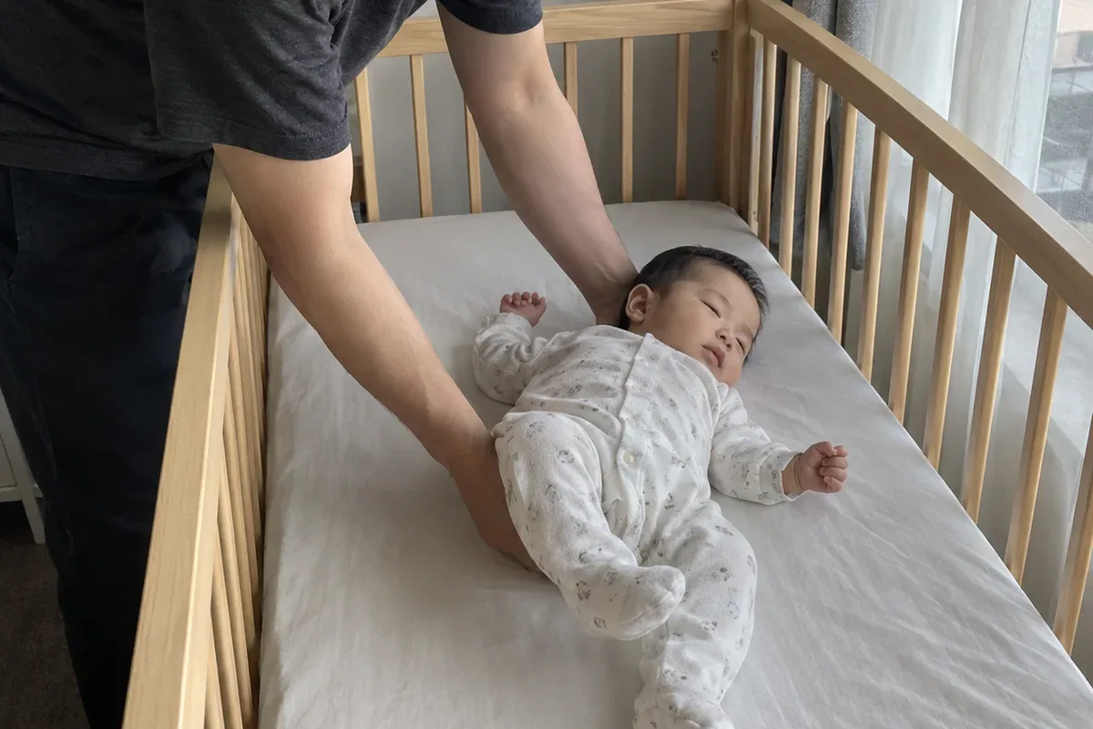 A parent's arms lowering a drowsy 9 month old onto their back in an empty crib with only a fitted sheet