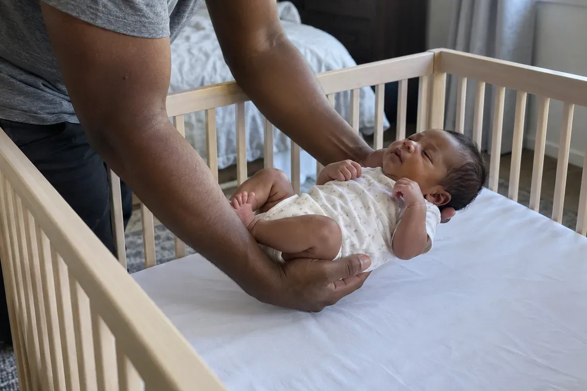 A parent's arms lowering a drowsy newborn onto their back in an empty crib with only a fitted sheet