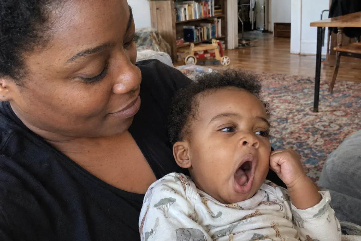 Close photo of a 1 year old in a parent's arms yawning widely and looking away, one fist near the face