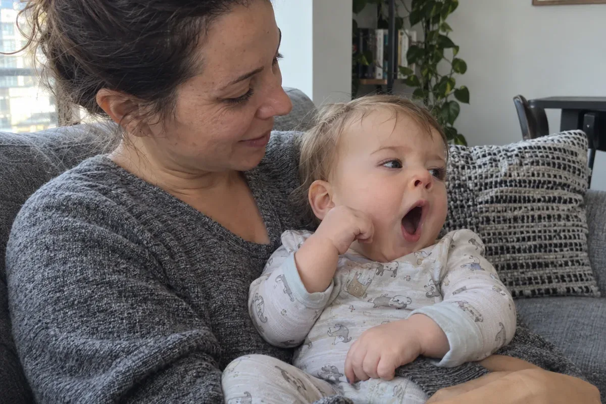 Close photo of a 10 month old in a parent's arms on the sofa mid-yawn, looking away with a fist near the face