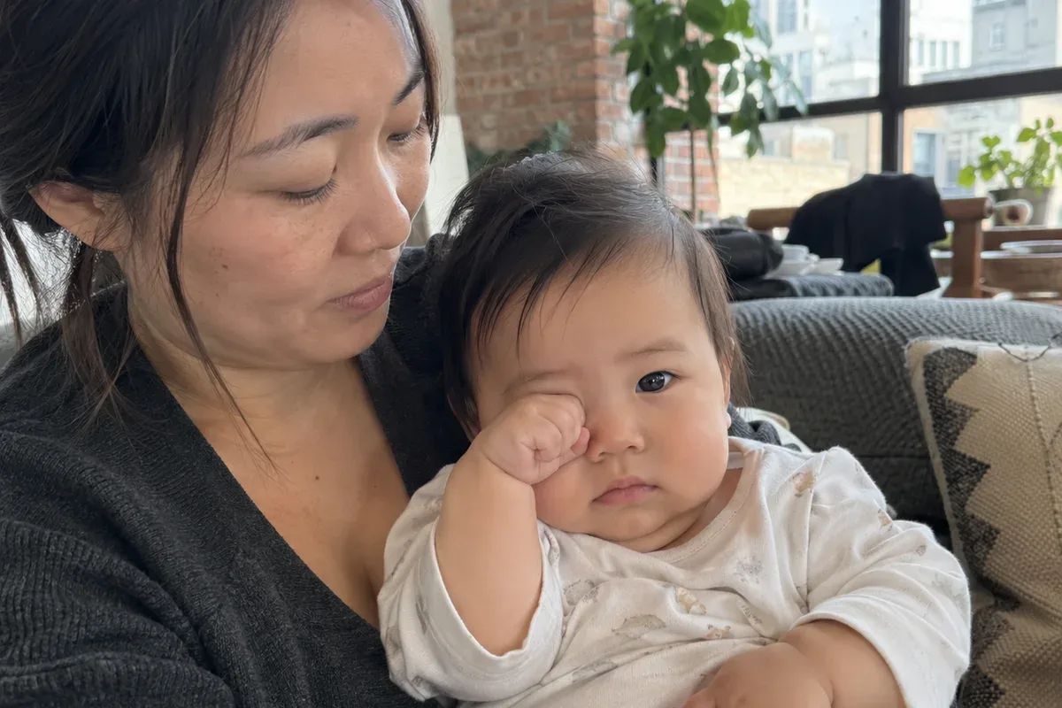 Close photo of a 9 month old in a parent's arms rubbing one eye with a fist and staring past the camera
