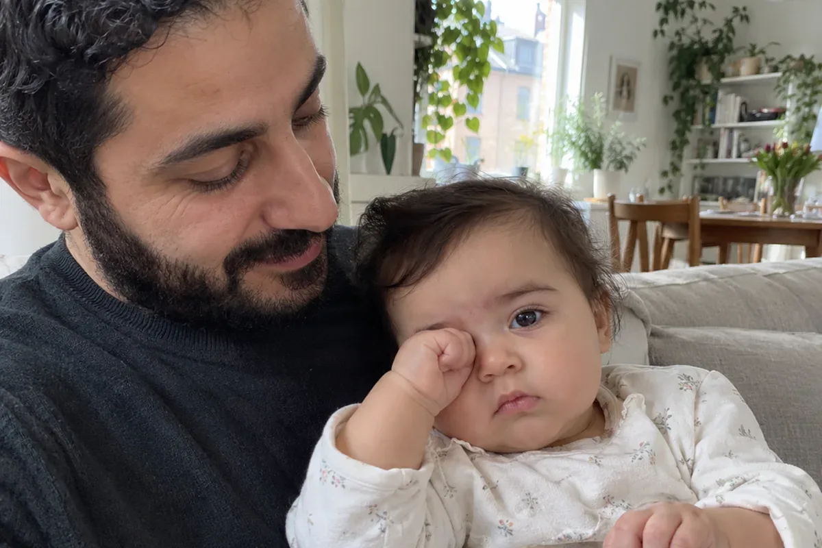 Close photo of an 11 month old in a parent's arms rubbing an eye with a fist and staring blankly past the camera