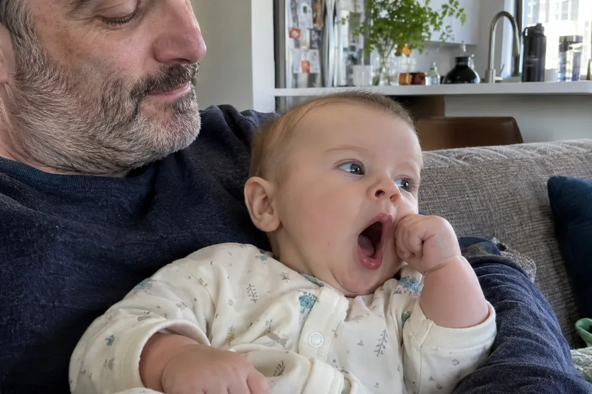 Close photo of an 8 month old in a parent's arms on the sofa, yawning and looking away with a fist near the face