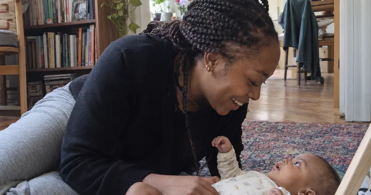 Parent kneeling beside a 6 week old during tummy time on a play mat in a daylit living room