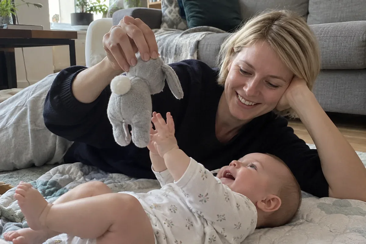 Parent kneeling on a play mat while a 4 month old does tummy time, pushing up on their forearms and reaching for a rattle
