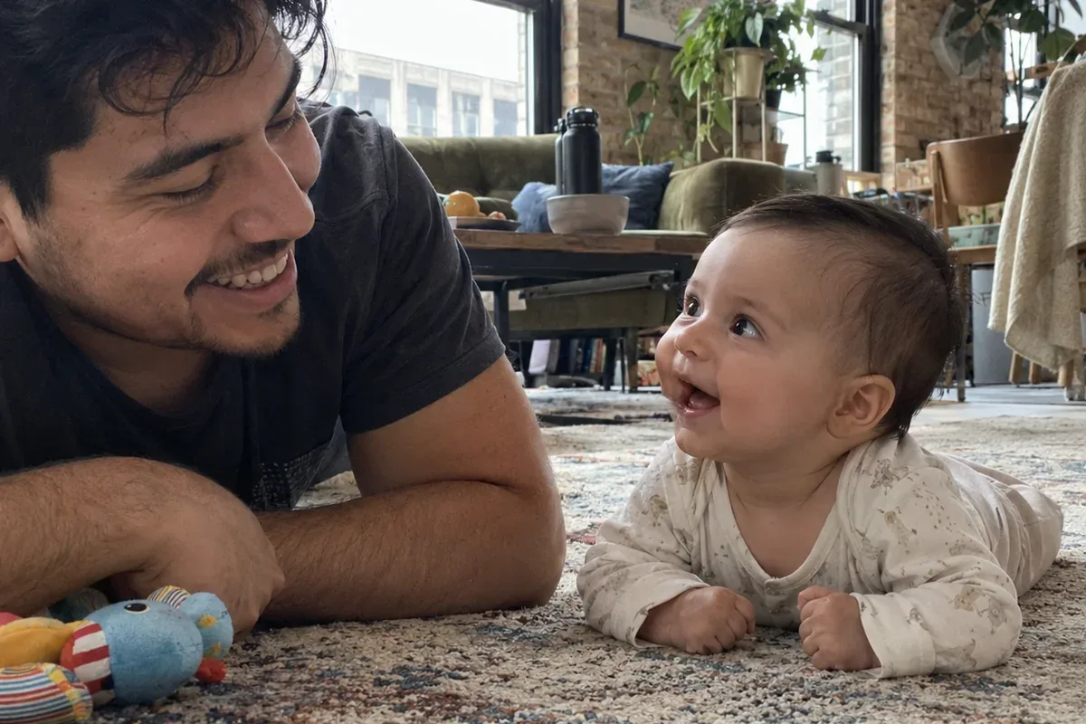Parent lying on a living room rug beside a 3 month old doing tummy time and reaching toward a soft toy