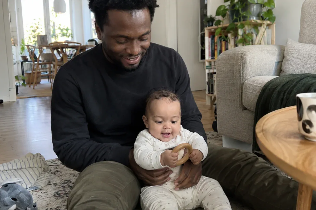 Parent sitting on the floor behind a 5 month old who sits with support and holds a stacking cup