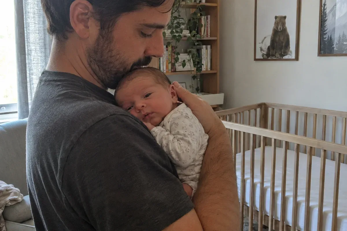 Parent swaying with a drowsy 3 week old on their shoulder in a dim nursery, empty bassinet nearby
