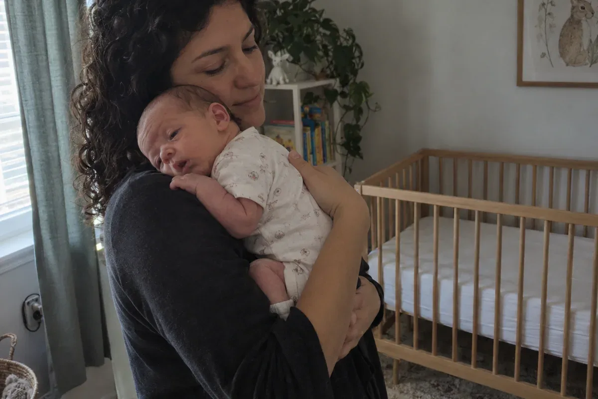 Parent swaying with a drowsy newborn on their shoulder in a dim nursery, empty crib in the background