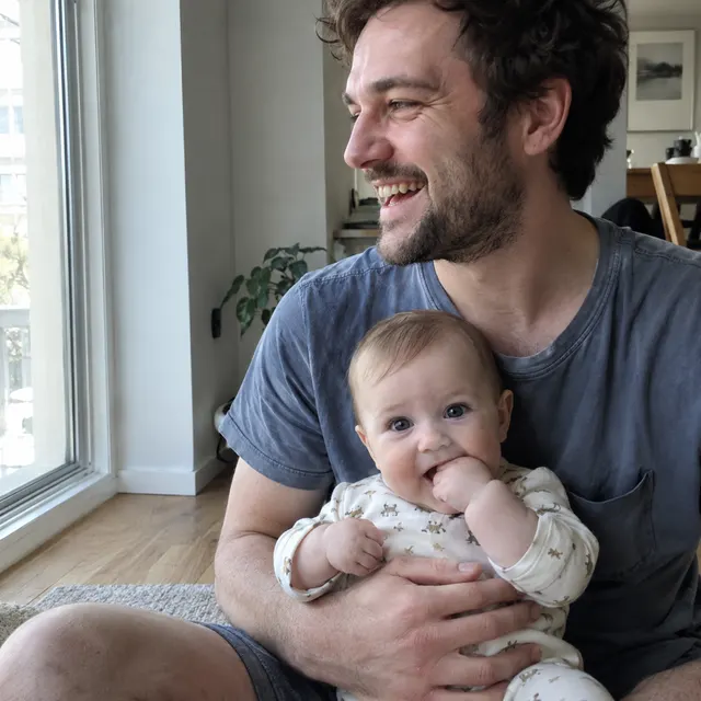 A father laughing on the floor by a window with his awake baby on his lap